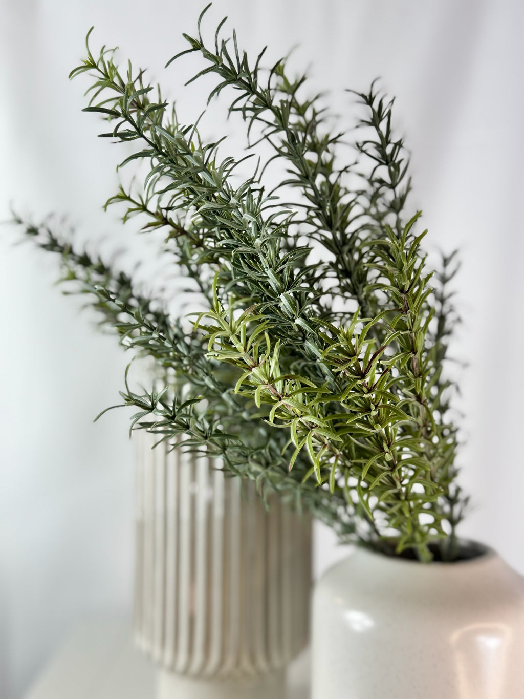 Artificial Bloom artificial rosemary botanical stem with realistic green needle-like leaves in a white ceramic vase.
