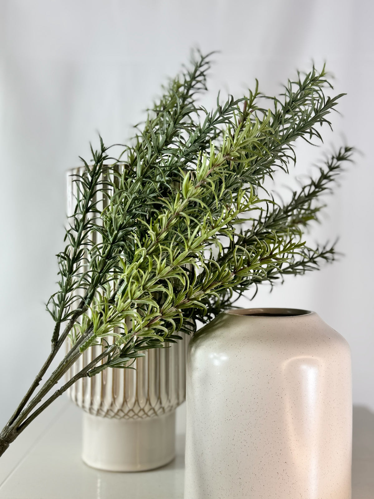 Artificial Bloom artificial rosemary botanical stems in a decorative white ceramic vase on a white surface