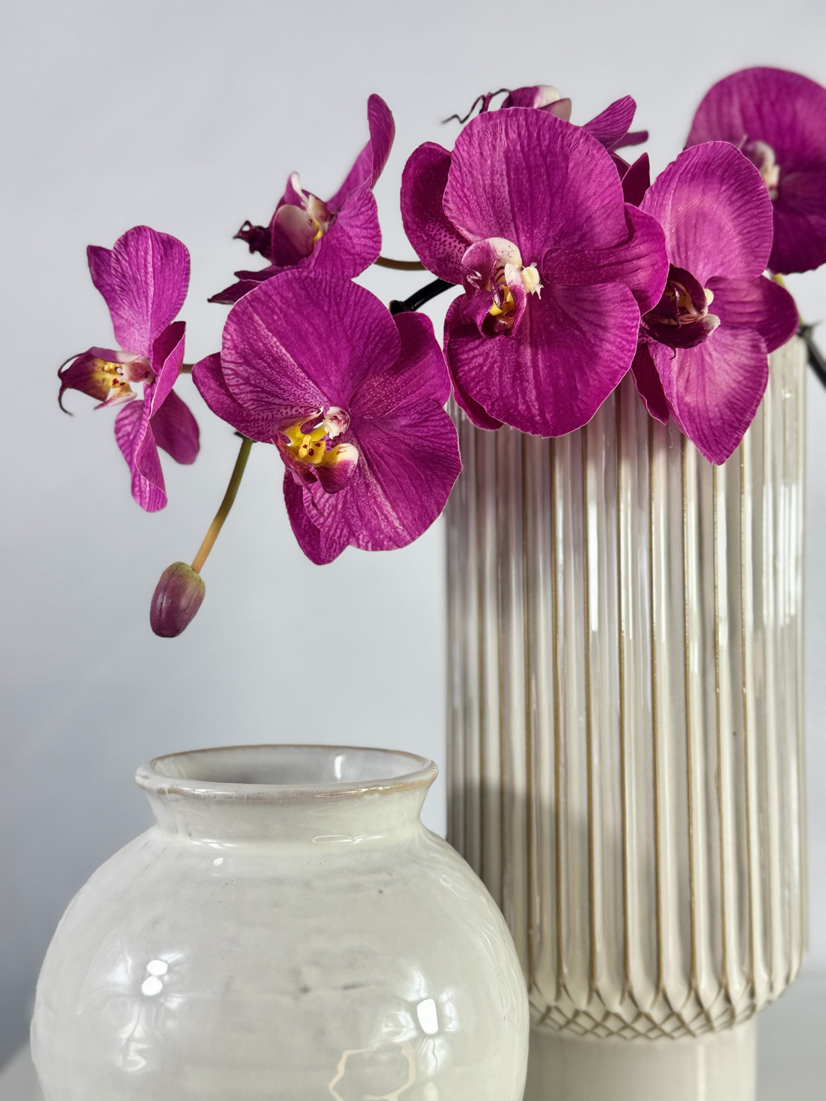 Artificial Bloom magenta Phalaenopsis orchid arrangement in a tall ribbed white ceramic vase next to a small bowl.