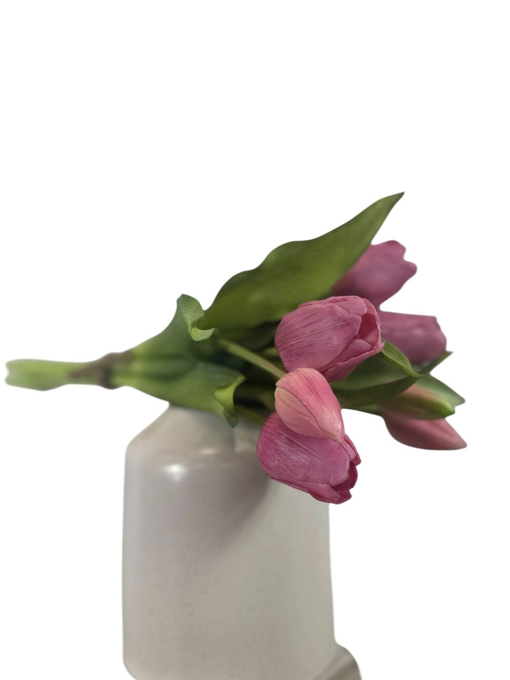 Artificial Bloom bouquet of realistic pink faux tulips with green foliage in a white vase against a white background.